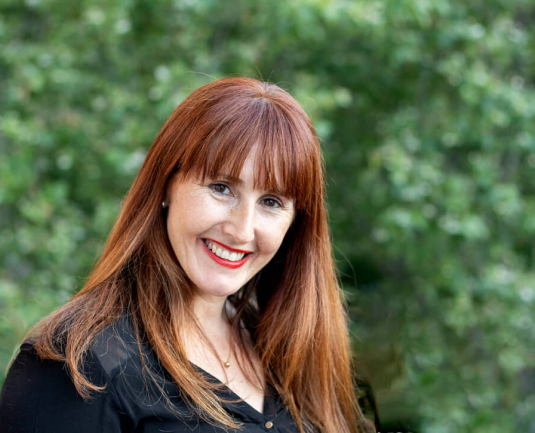 Eva Ortiz, copywriter freelance, sonríe a cámara, de fondo unos árboles.